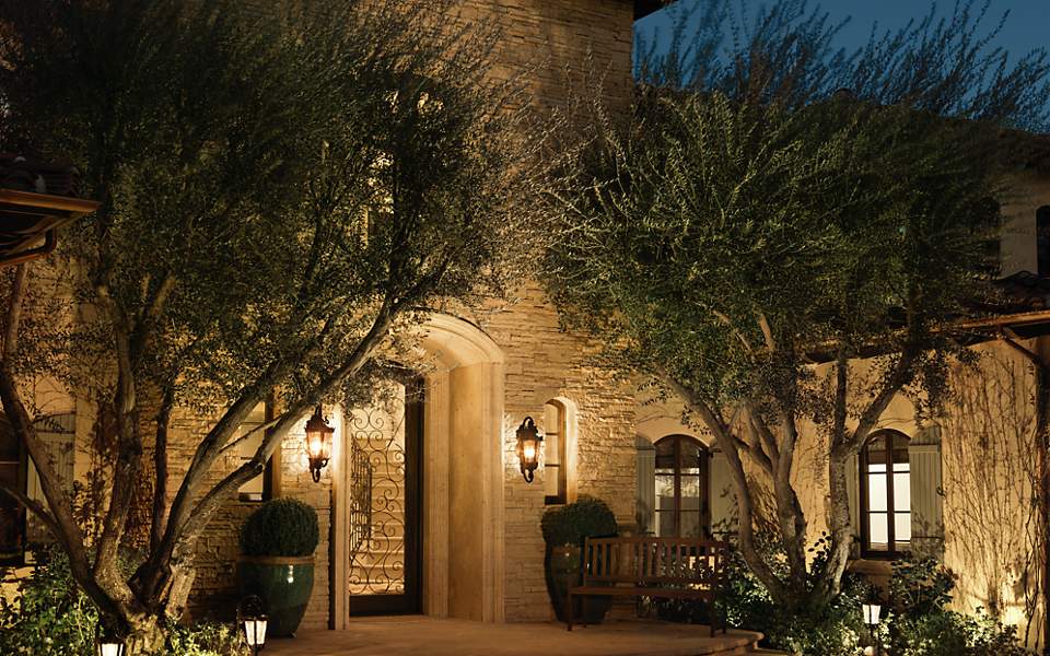 Outdoor house scene with bright lighting, including two bronze wall lights. Spanish-colonial house that glows brightly at night due to the lighting, with two outdoor wall lights visible.
