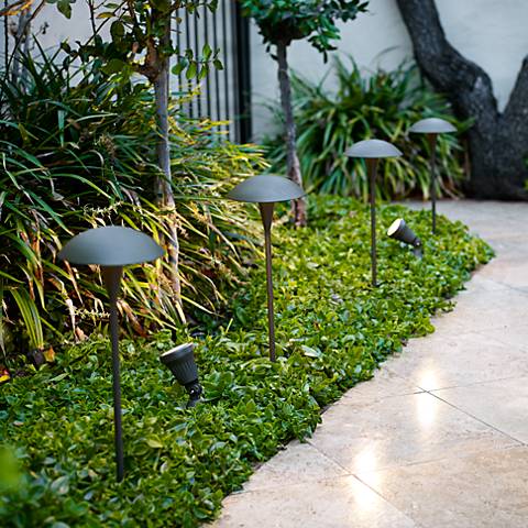 A series of bronze mushroom path lights, set in a green garden next to a stone path. Four dark bronze mushroom-shaped path lights.