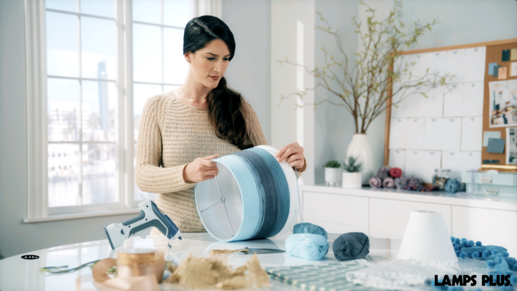 A woman decorating a lamp shade with yarn. 